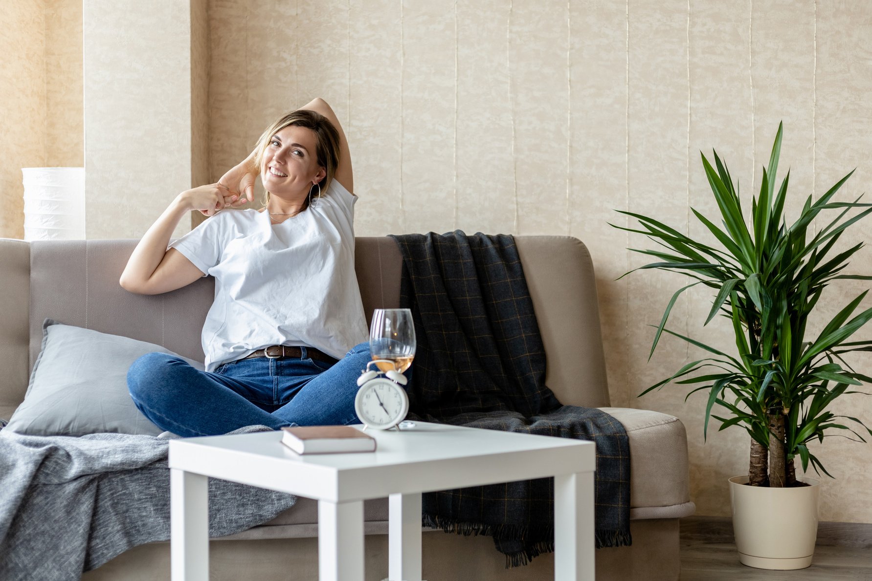 young smiling blonde girl in a white t-shirt and jeans sits on a sofa in a bright room